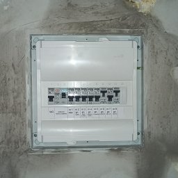PAWEŁ OPALIŃSKI OPAL ELEKTRO MONTAŻ I POMIARY INSTALACJI ELEKTRYCZNYCH - Zamknięta skrzynka rozdzielcza z bezpiecznikami w szarej obudowie, zamontowana w ścianie z widocznymi śladami po remoncie. Widoczne opisy bezpieczników: salon, łazienka, zmywarka, piekarnik, piec...