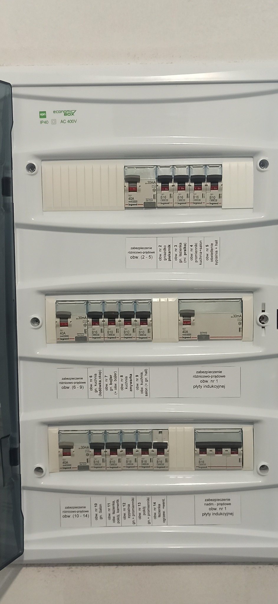 Rozdzielnica elektryczna z widocznymi bezpiecznikami różnicowoprądowymi i nadprądowymi firmy Legrand, oznaczonymi obwodami dla piekarnika, łazienki, kuchni, salonu, sypialni oraz płyty indukcyjnej.
