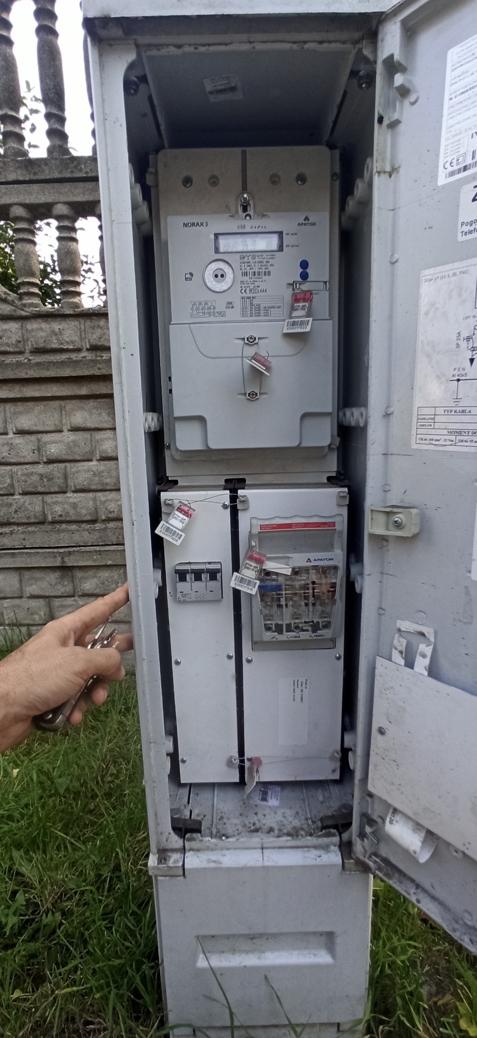 Otwarta skrzynka elektryczna z licznikiem i bezpiecznikami, plombami, w tle mur z cegły i trawa, ręka z kluczem.