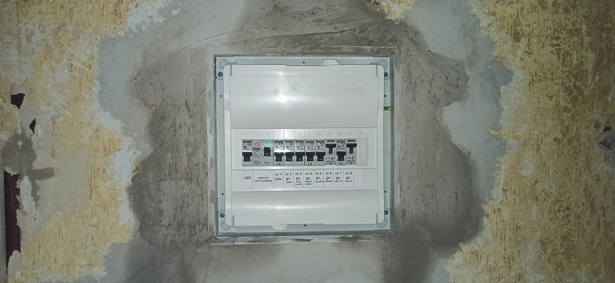 Zamknięta skrzynka rozdzielcza z bezpiecznikami w szarej obudowie, zamontowana w ścianie z widocznymi śladami po remoncie. Widoczne opisy bezpieczników: salon, łazienka, zmywarka, piekarnik, piec...