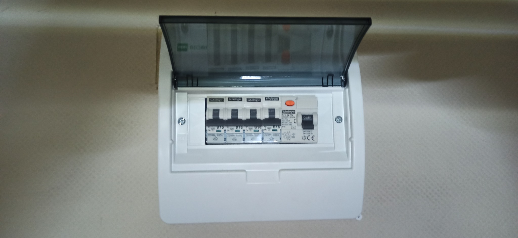 Otwarta skrzynka elektryczna z bezpiecznikami marki Schaltinger, zamontowana na ścianie o jasnej strukturze.