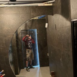 Hubert Samulak - Ciemne płytki w łazience z odbiciem osoby robiącej zdjęcie w okrągłym lustrze. Widoczne rury pod sufitem i elementy montażowe.