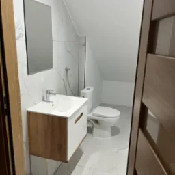 Nowoczesna łazienka na poddaszu z biało-marmurowymi płytkami, drewnianą szafką pod umywalką i lustrem bez ramy. Widok zza uchylonych drzwi.