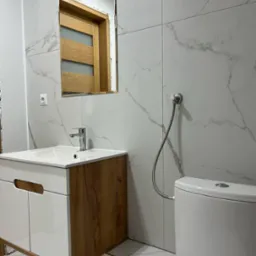 Nowoczesna łazienka z marmurowymi płytkami, szafką pod umywalką z drewnianymi akcentami i lustrem odbijającym drzwi. Minimalistyczny design i oświetlenie.