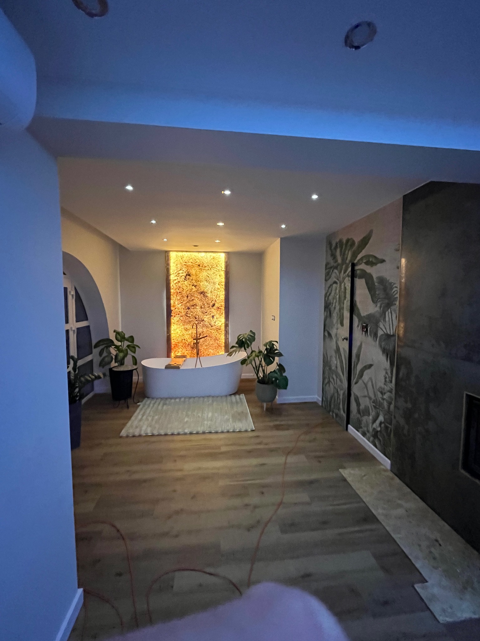 Eklektyczna łazienka z wolnostojącą wanną na tle podświetlanej ściany i egzotycznej tapety, uzupełniona roślinami doniczkowymi i drewnianą podłogą.