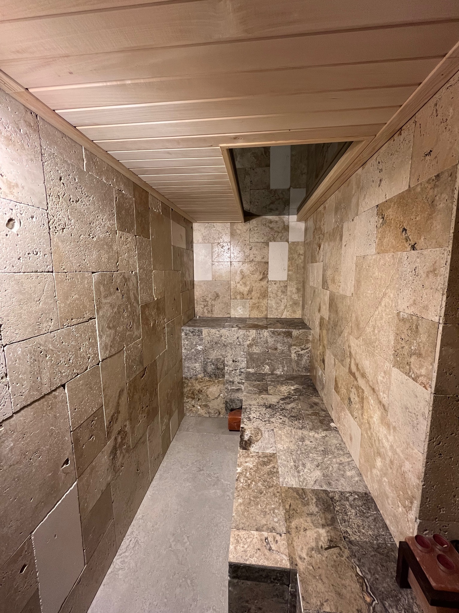 Wnętrze sauny fińskiej wyłożone trawertynem. Widoczne drewniane panele sufitowe i szklany świetlik. Kamienne ławy i podłoga z płytek w odcieniach beżu i brązu.