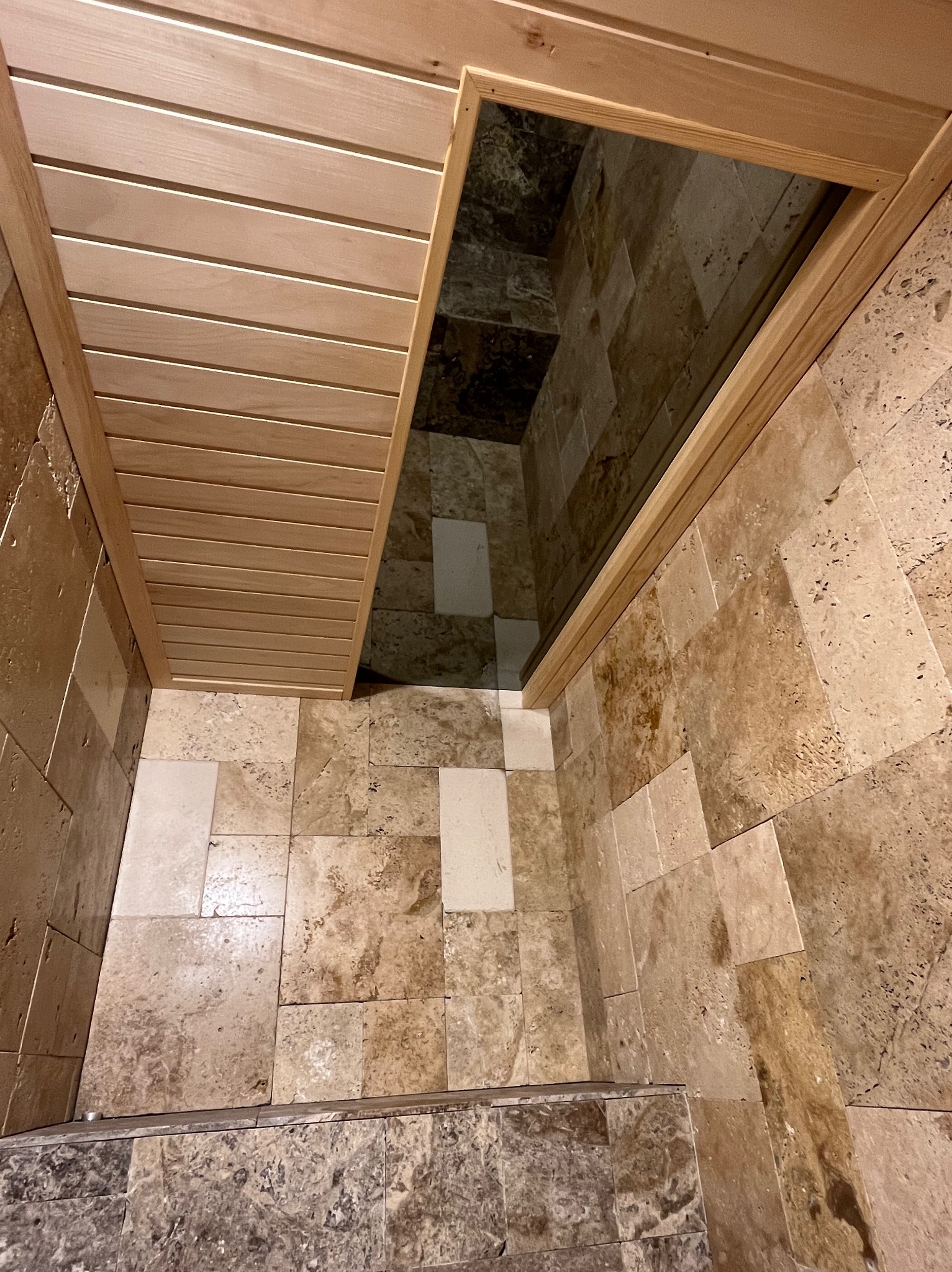 Wykończona łazienka z beżowymi płytkami trawertynowymi na podłodze i ścianach, drewnianym sufitem i wejściem do kabiny prysznicowej.