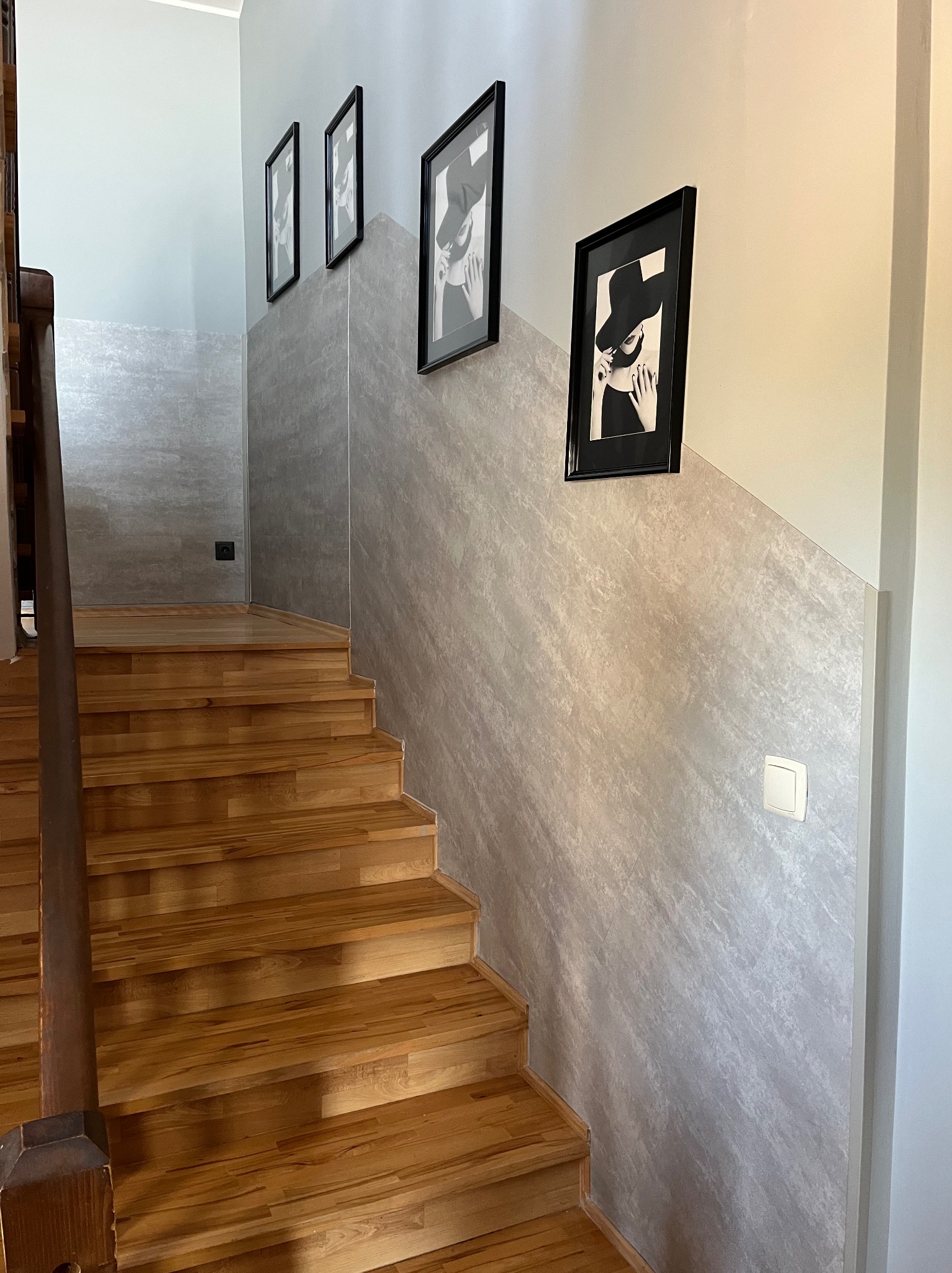 Drewniane schody z balustradą, ściana wykończona w szarości, z trzema obrazami w czarnych ramkach. Widoczny włącznik światła i gniazdko elektryczne.
