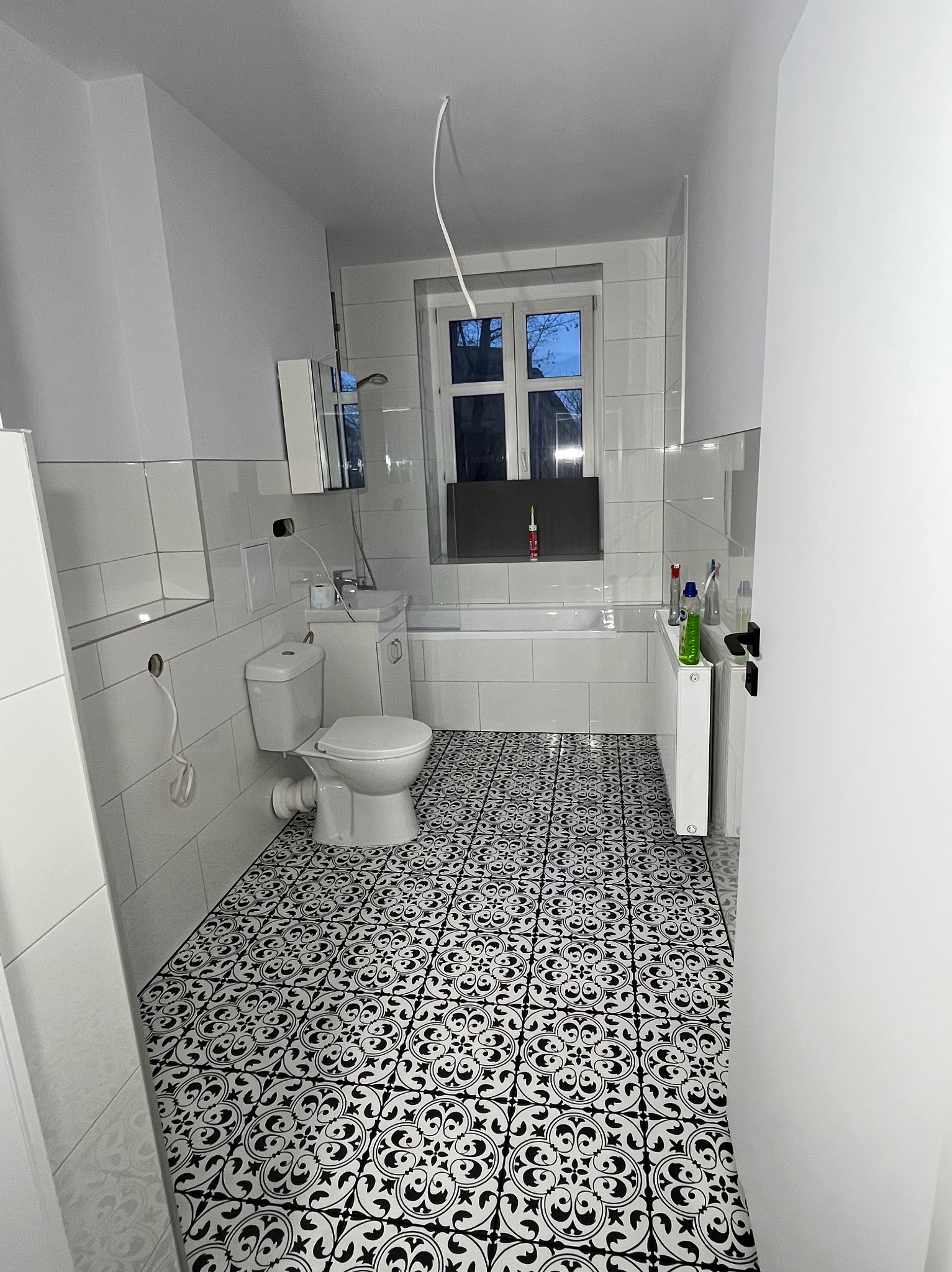 Biała łazienka z wanną, umywalką i toaletą. Na podłodze czarno-białe płytki w marokański wzór. Widoczne okno z fragmentem nieba.