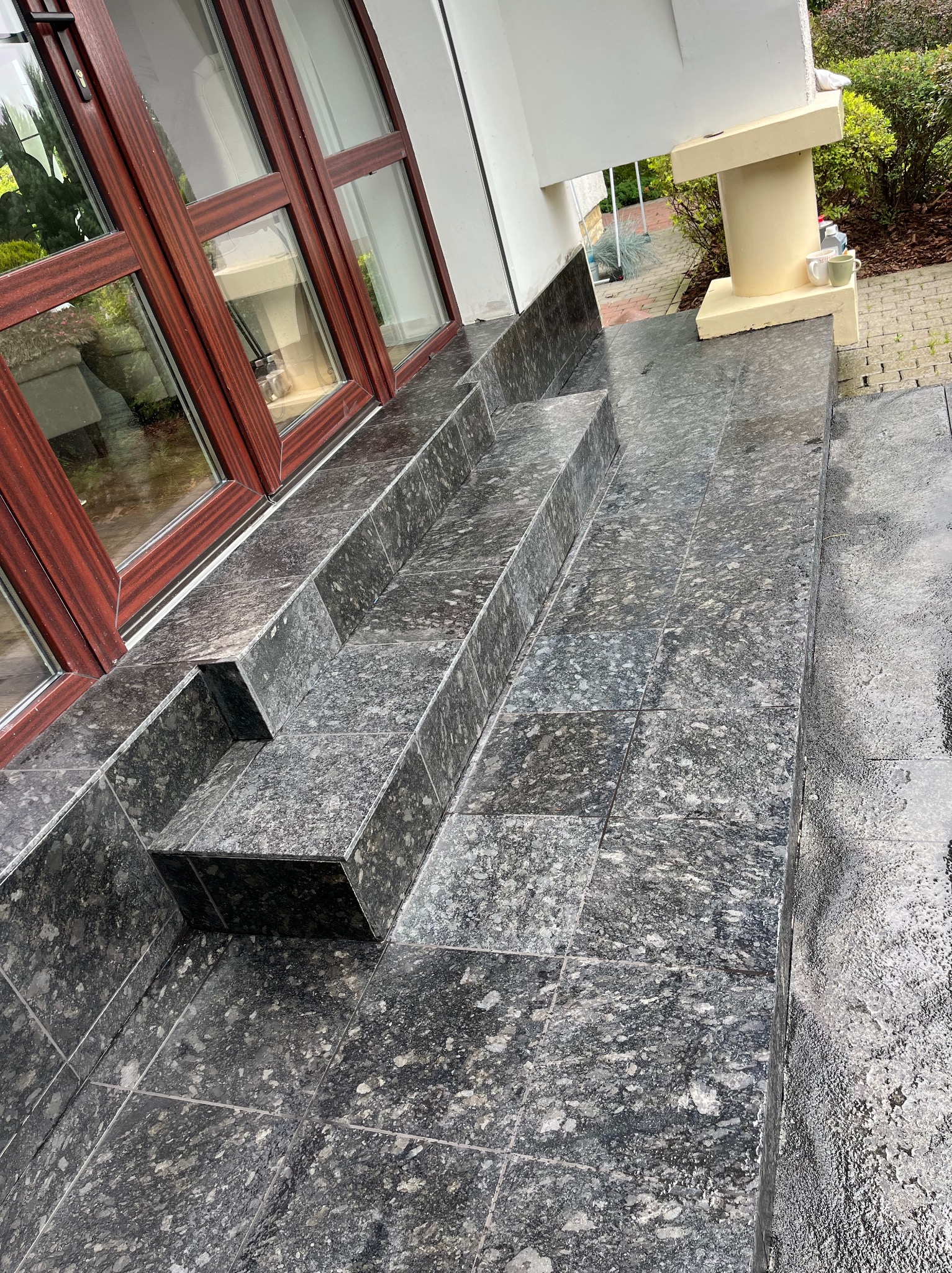 Granitowe schody zewnętrzne z ciemnoszarą, nakrapianą płytką, prowadzące do wejścia z brązowymi ramami okiennymi. Widok z góry, lekko z boku, na mokrą powierzchnię.