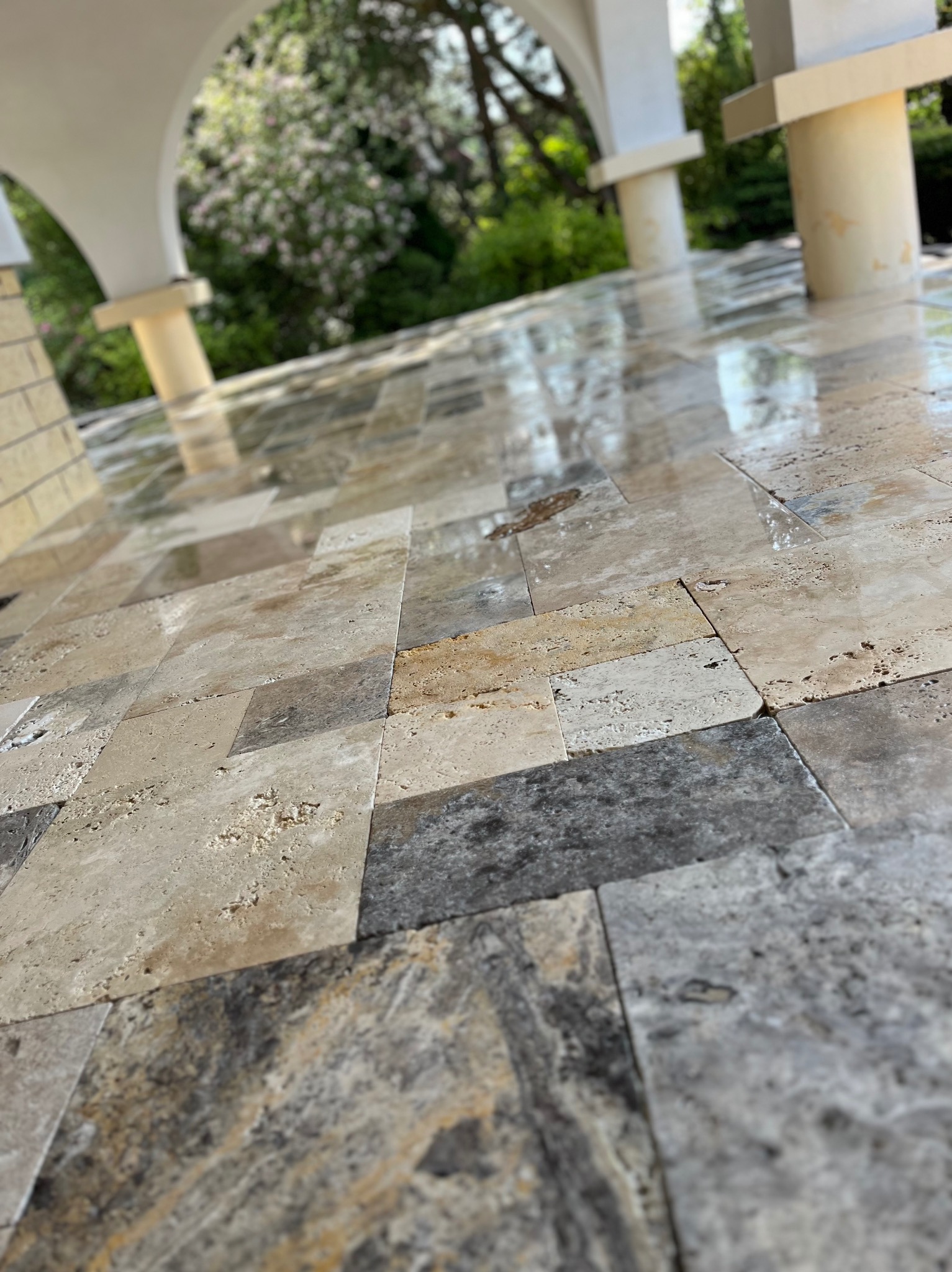 Błyszcząca posadzka z trawertynu w odcieniach beżu i szarości, ułożona w kwadraty, pod zadaszeniem z kolumnami. Widok z bliska, podkreślający fakturę kamienia.