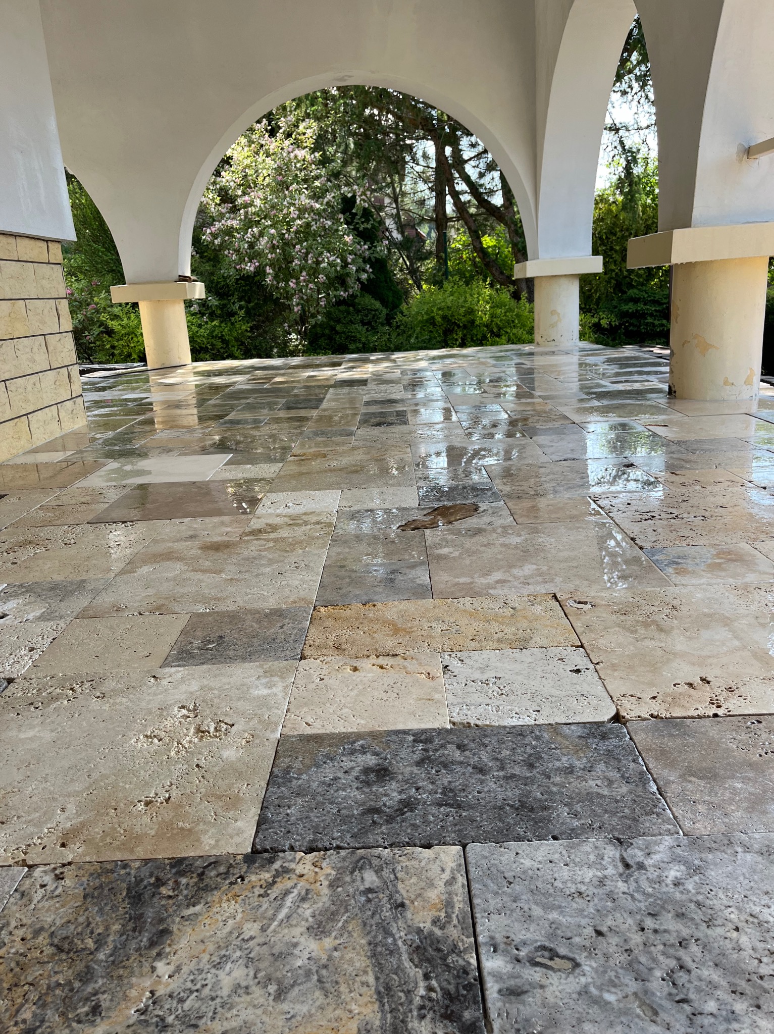 Położona, błyszcząca pod wpływem światła podłoga z trawertynu na zewnątrz budynku z arkadami i zielenią w tle. Widok na ułożone płyty z bliska.
