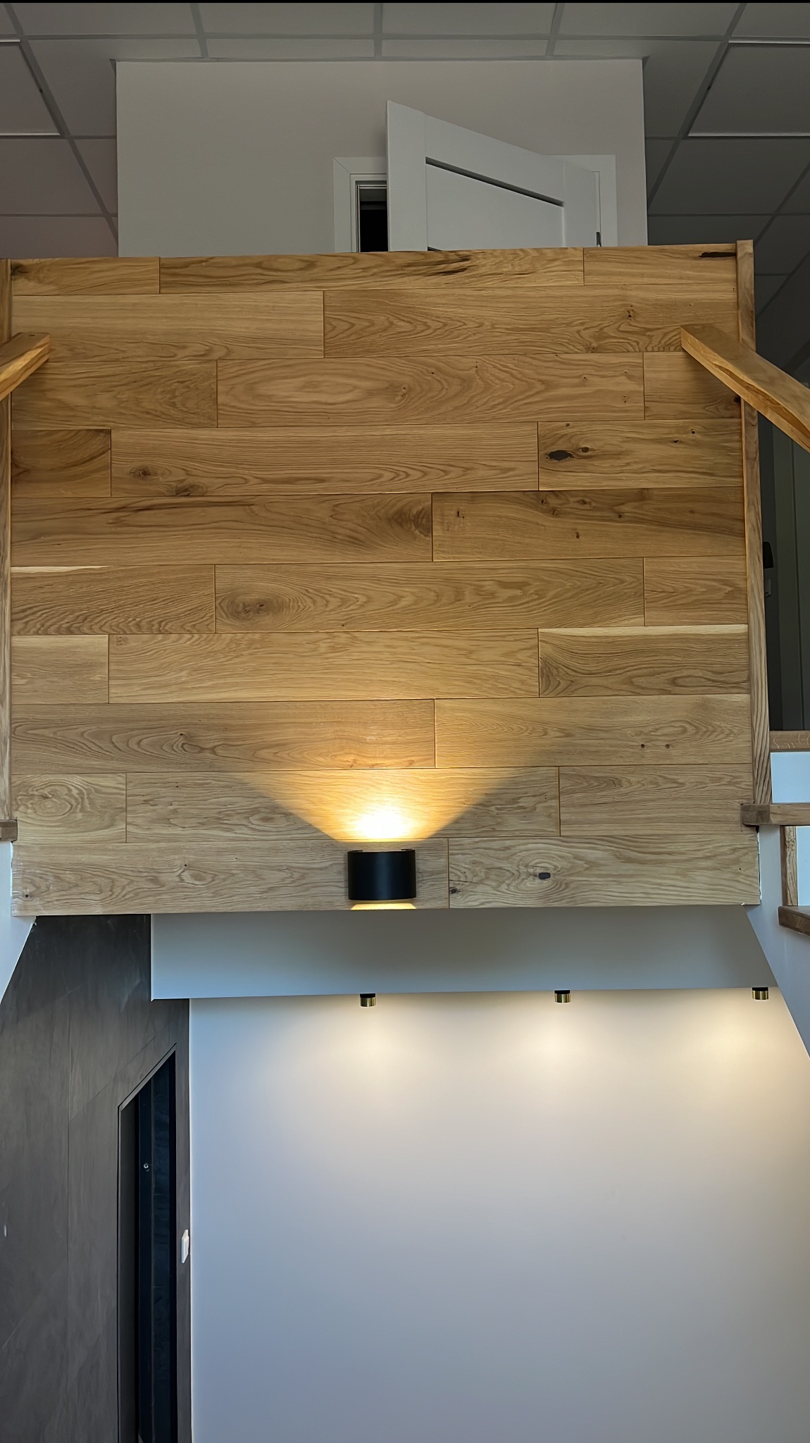 Drewniana ściana z poziomych paneli o ciepłym odcieniu, oświetlona przez czarny kinkiet, fragment schodów i biała ściana z trzema punktami świetlnymi.