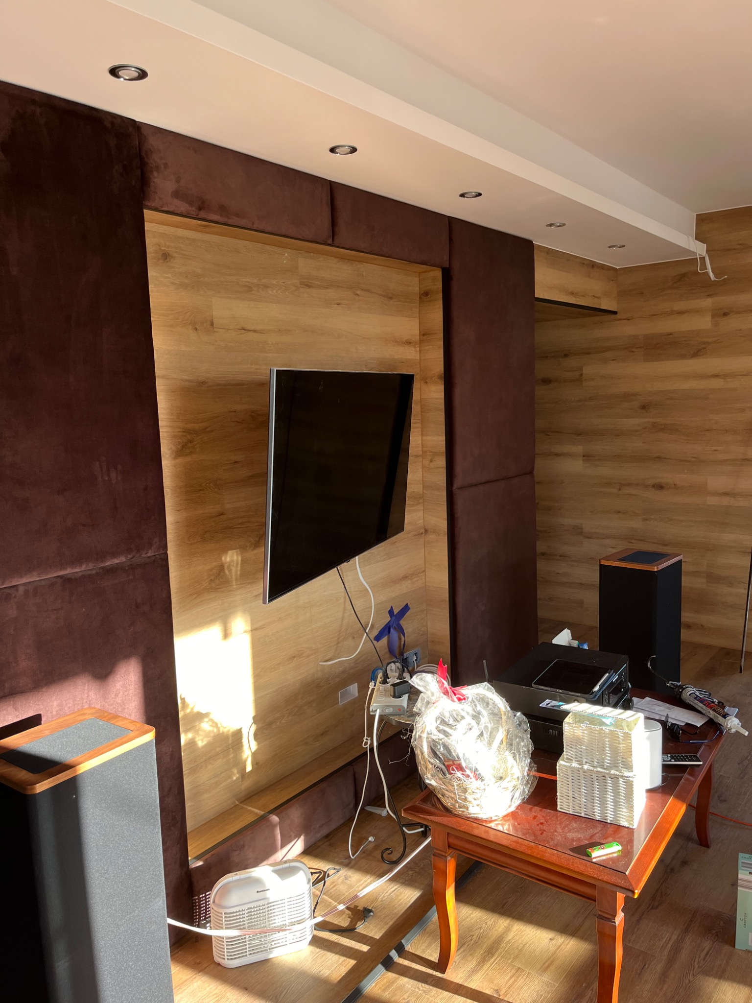 Wykończenie ściany z drewnianym panelem pod telewizor, obok brązowe panele tapicerowane. Na podłodze głośnik i grzejnik, na stole akcesoria i prezenty.