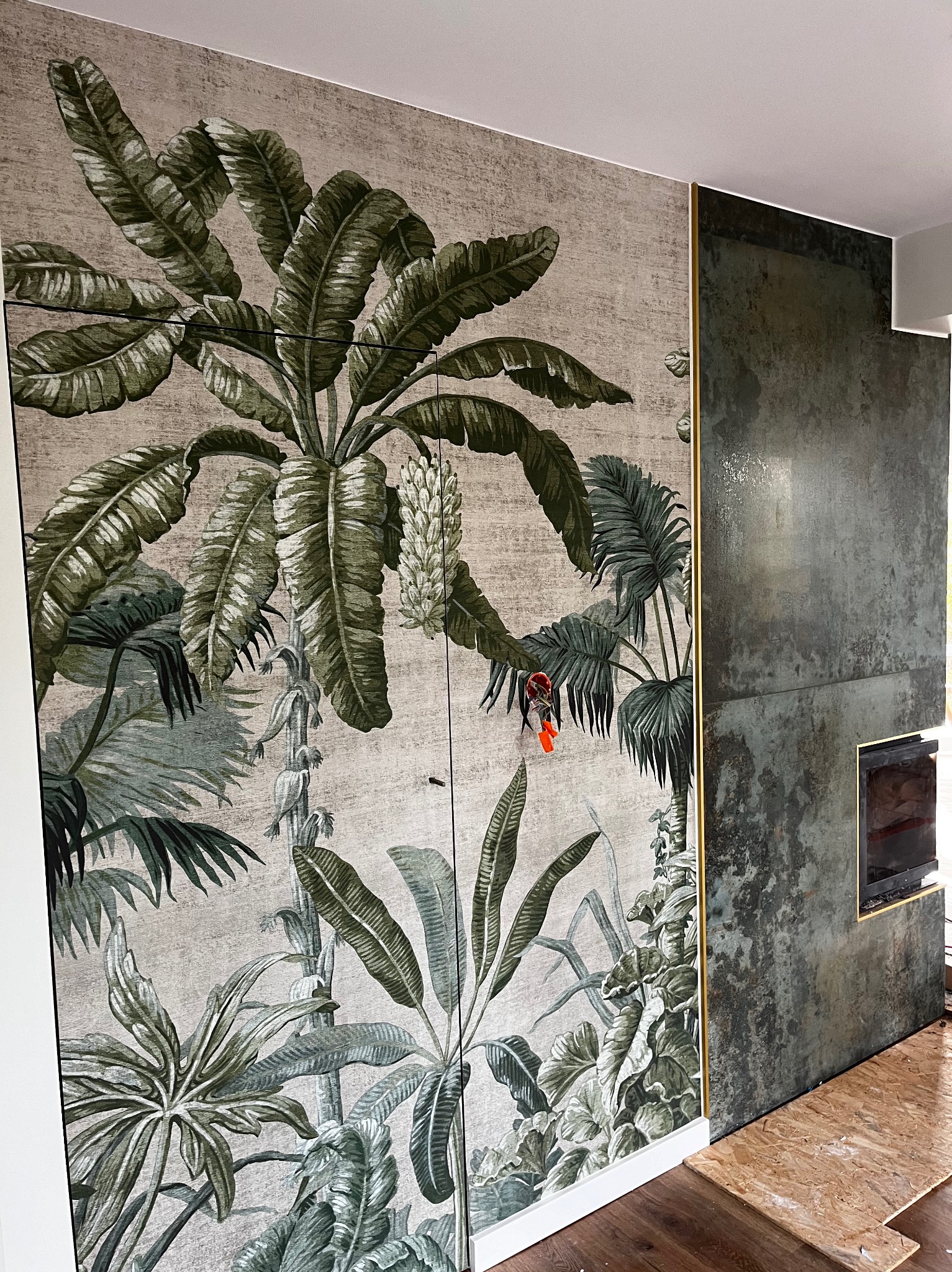 Egzotyczna tapeta z motywem roślin tropikalnych na ścianie, obok fragmentu ściany wykończonej płytami imitującymi metal, z widocznym kominkiem i fragmentem podłogi.