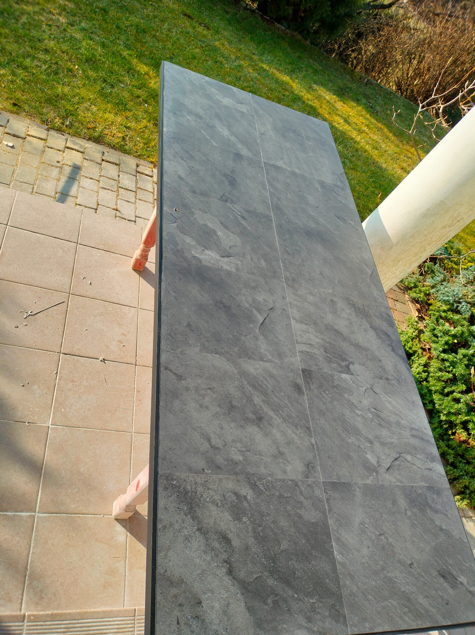 Szara, kamienna posadzka tarasu z widocznymi fugami, w tle trawnik i roślinność. Kąt ujęcia z góry podkreśla fakturę płytek i ich regularny układ.