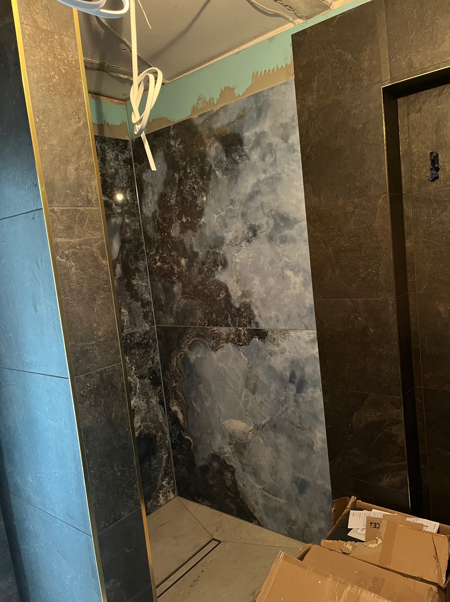 Fragment łazienki w trakcie wykańczania: marmurowe płytki w odcieniach szarości i błękitu na ścianie, obok ciemne płytki z złotymi listwami. Widoczne kartony na podłodze.