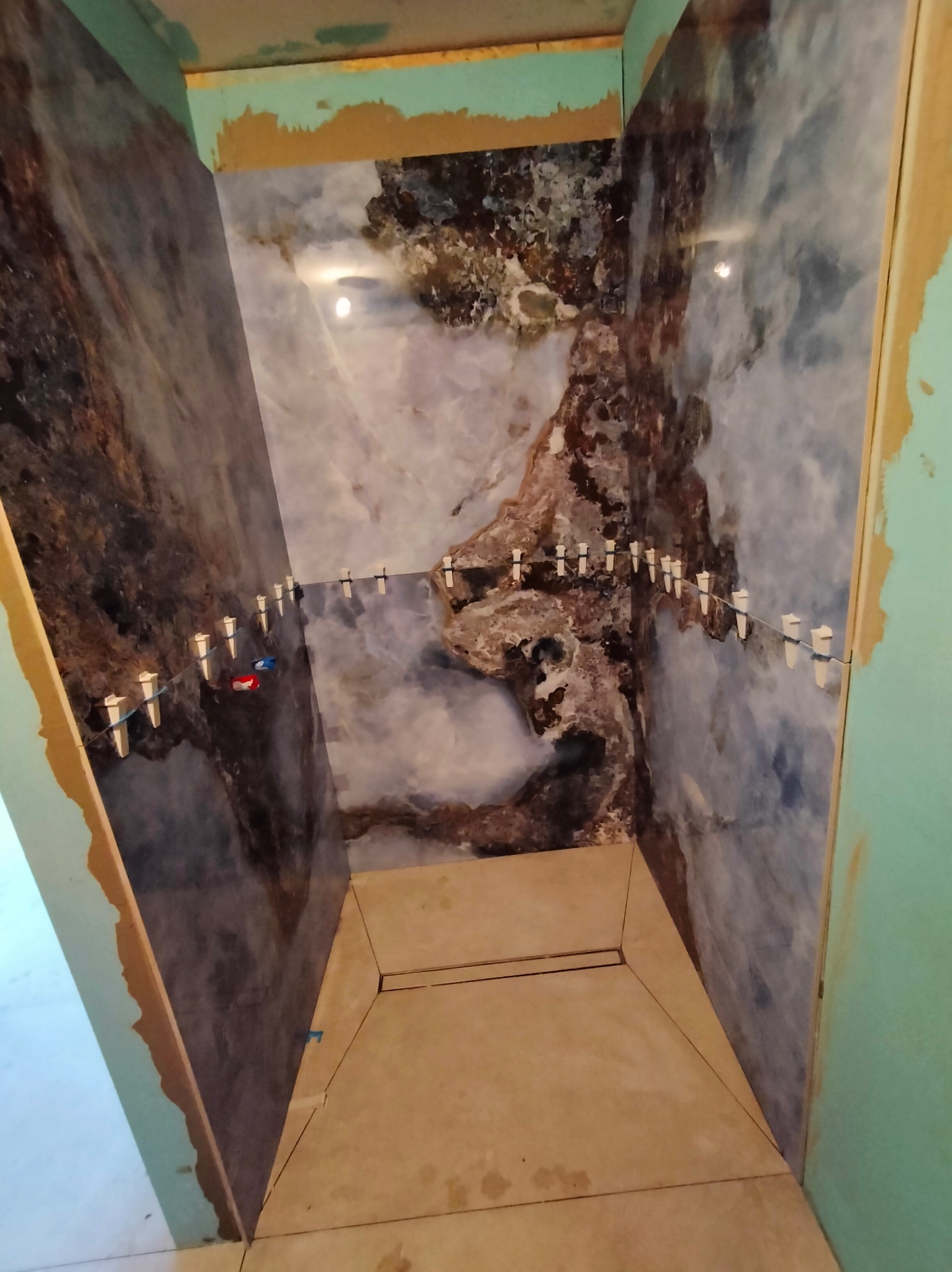 Montaż płytek imitujących marmur w kabinie prysznicowej, z użyciem systemu poziomowania. Widoczne płytki na ścianach i podłodze, zielone płyty g-k na krawędziach.