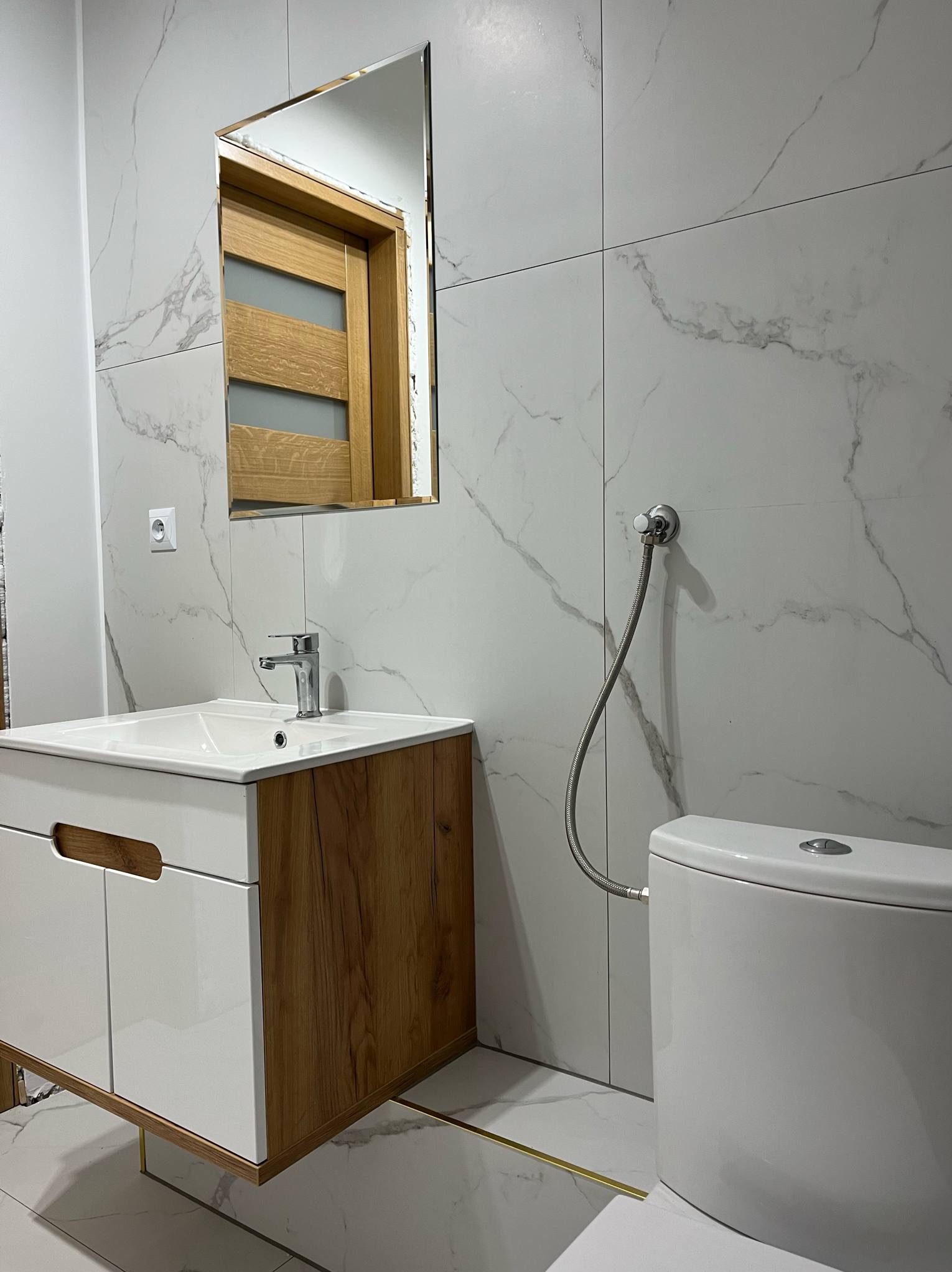 Nowoczesna łazienka z marmurowymi płytkami, szafką pod umywalką z drewnianymi akcentami i lustrem odbijającym drzwi. Minimalistyczny design i oświetlenie.