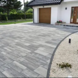 Nowoczesny podjazd z szarej kostki o nieregularnym wzorze, obrzeże z ciemnej kostki, biały żwir, dom z brązowymi drzwiami garażowymi i wejściowymi w tle.