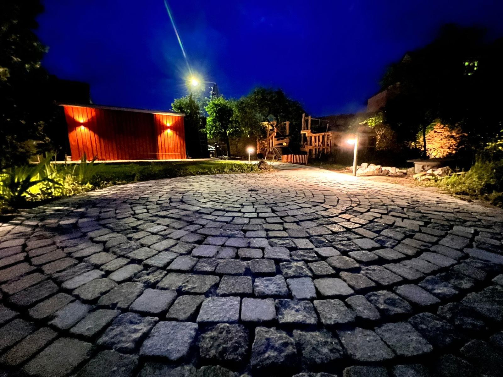 Nocny widok na okrągły plac wyłożony szarą kostką granitową w Zielonej Górze, oświetlony lampami, z drewnianym domkiem w tle.