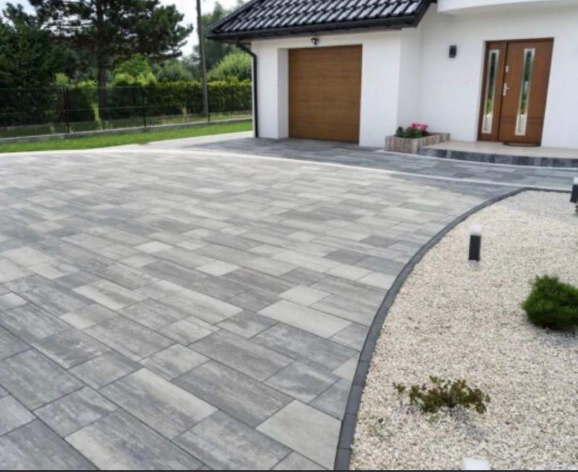 Nowoczesny podjazd z szarej kostki o nieregularnym wzorze, obrzeże z ciemnej kostki, biały żwir, dom z brązowymi drzwiami garażowymi i wejściowymi w tle.