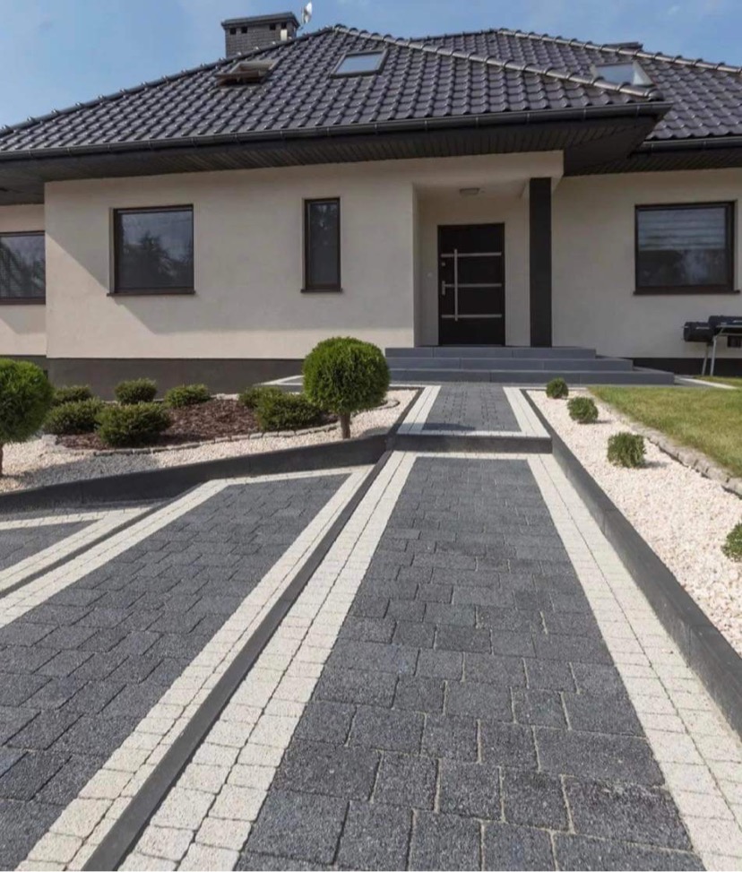 Nowoczesny podjazd do domu wyłożony szarą kostką z białymi obrzeżami, z elementami małej architektury i zadbanym trawnikiem.