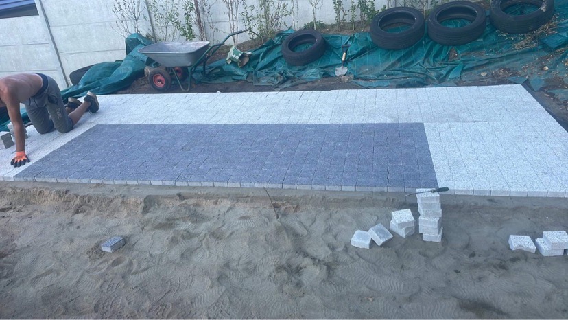 Prace brukarskie w toku: robotnik układa mozaikę z kostki granitowej w dwóch odcieniach, jasnoszarym i ciemnoszarym, widoczny stos kostek, taczka i opony w tle.