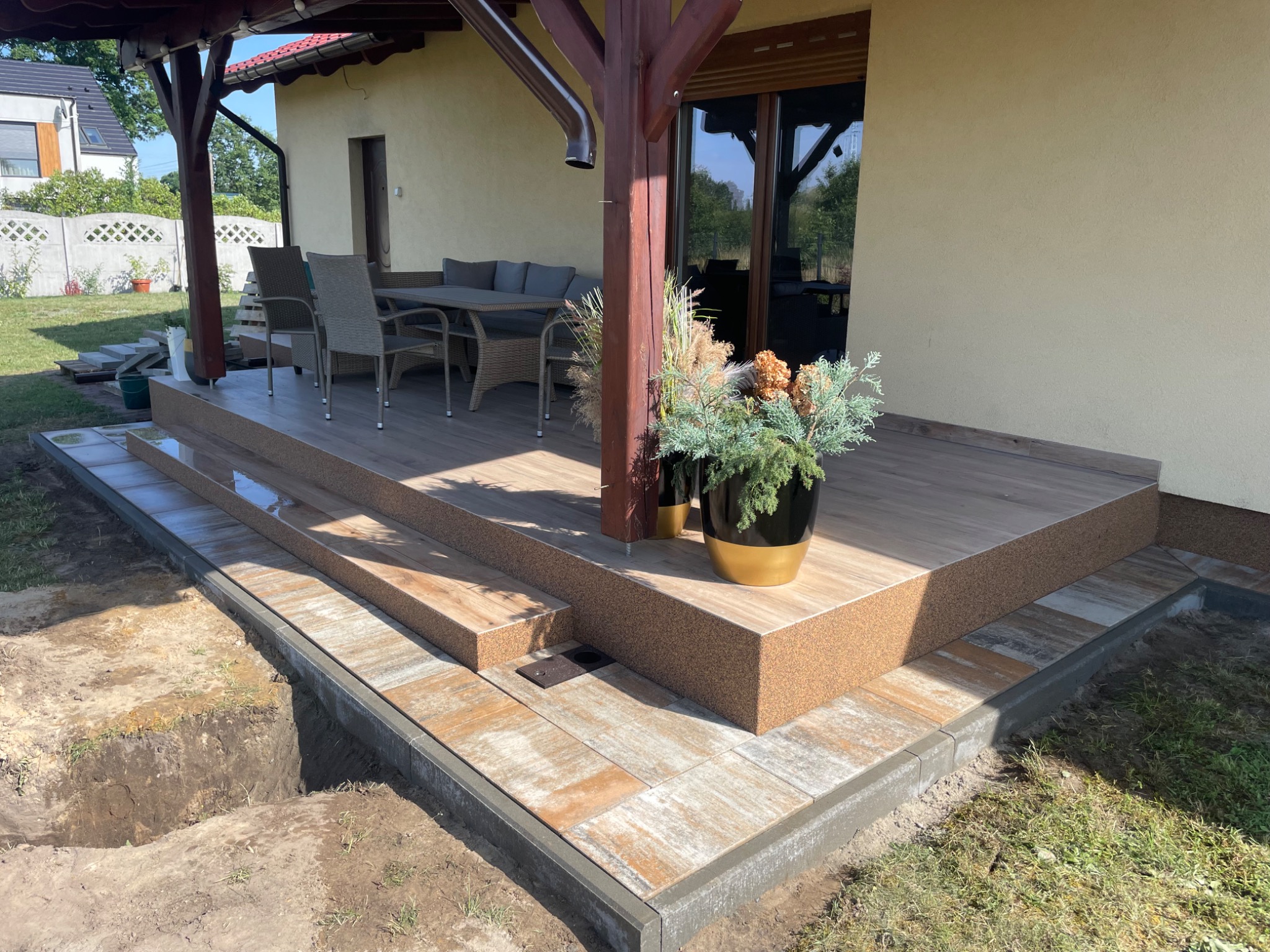 Wykończony taras z kostki brukowej w odcieniach beżu i brązu, z widocznymi schodami i donicami z roślinami, obok wykop pod dalszą część tarasu.