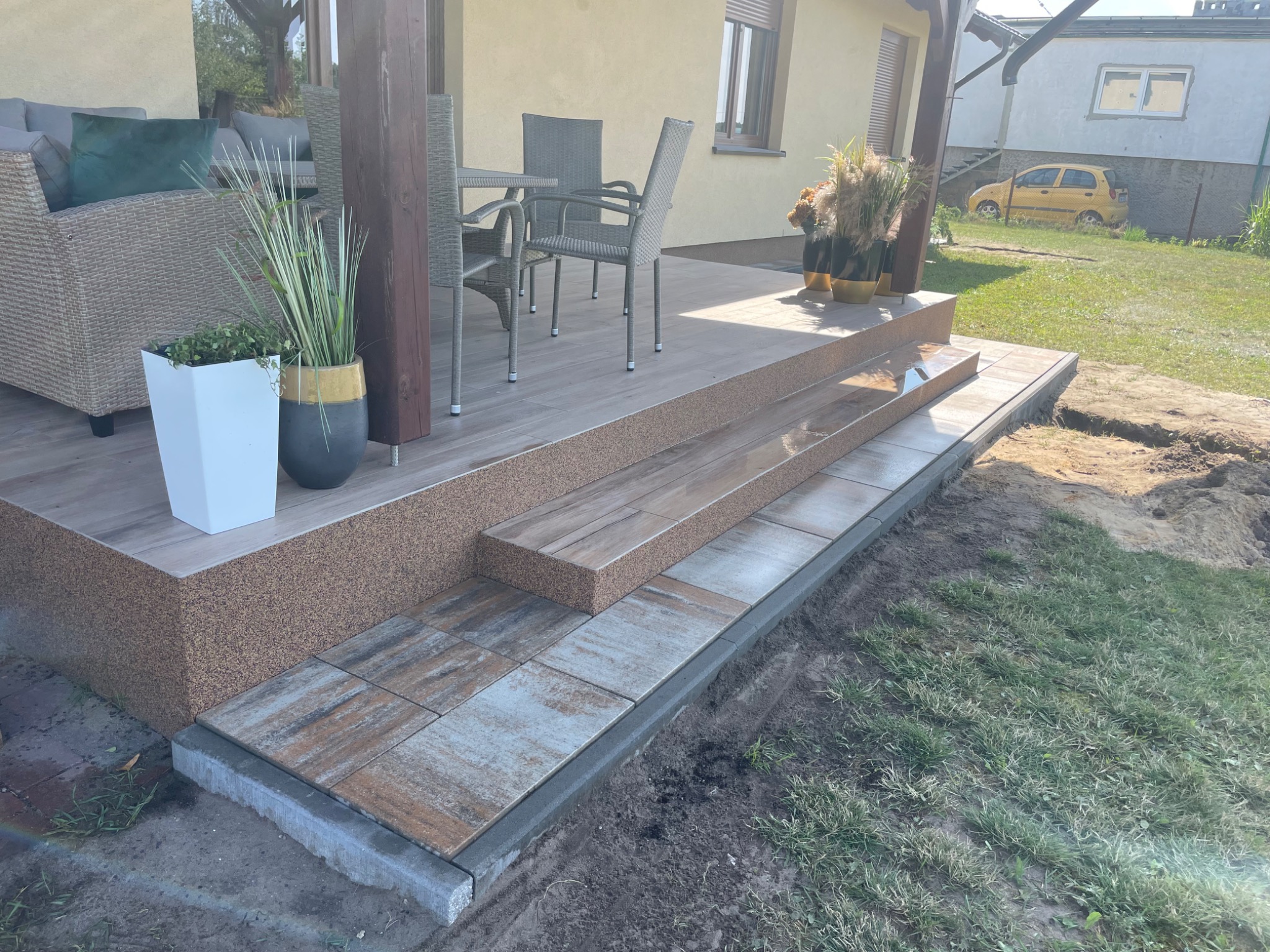 Wykończony taras z drewnopodobną podłogą i schodami wyłożonymi płytkami na zewnątrz domu, widoczne meble ogrodowe i rośliny w donicach, w tle zaparkowany żółty samochód.