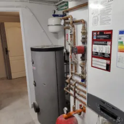 Szary zbiornik akumulacyjny obok białego kotła gazowego Ariston z licznymi miedzianymi rurami i zaworami w kotłowni.