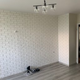 Simplex - Pokój po remoncie z białą ścianą wyłożoną dekoracyjną cegłą, szarymi panelami podłogowymi i nowoczesną lampą sufitową z kloszami w kształcie kul. Przy ścianie leżą przewody i urządzenia elektroniczne.