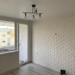 Simplex - Pusty pokój po remoncie: biała ceglana ściana, szara podłoga z paneli, nowoczesna lampa sufitowa z czterema szklanymi kloszami, okno balkonowe z widokiem na blok, kable elektryczne zwisające ze...