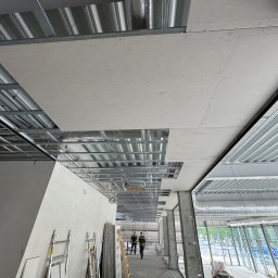 Simplex - Montaż sufitu podwieszanego z płyt karton-gips na metalowej konstrukcji. Widoczne płyty, profile, drabiny i pracownicy w oddali. Etap prac wykończeniowych w budynku.