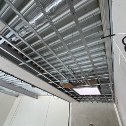 Simplex - Metalowa konstrukcja sufitu podwieszanego z widoczną izolacją, instalacją elektryczną i elementami oświetlenia w surowym wnętrzu. Widok z dołu.