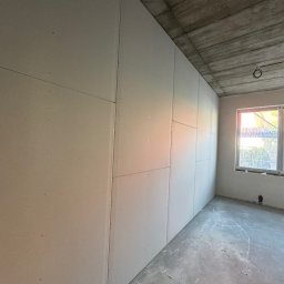 Simplex - Montaż płyt na ścianie w surowym wnętrzu w Warszawie. Widoczne łączenia płyt i betonowy sufit. Okno z widokiem na zieleń.