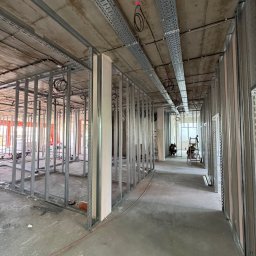 Simplex - Wnętrze w trakcie prac: metalowa konstrukcja ścian działowych, koryta kablowe pod sufitem, surowy beton, widoczni pracownicy na drabinie w głębi korytarza.