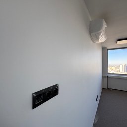 Simplex - Świeżo pomalowana ściana w biurze z gniazdkami elektrycznymi, klimatyzacja w folii ochronnej i widokiem na miasto z okna. Jasne wnętrze po odnowieniu.