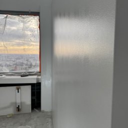 Simplex - Świeżo pomalowana, gładka ściana w odcieniach szarości w pokoju z widokiem na miasto, z wiaderkiem farby i wałkiem na podłodze. Okno zabezpieczone folią malarską.