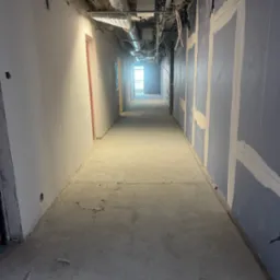 Długi korytarz w trakcie prac wykończeniowych. Widoczne instalacje pod sufitem, ściany przygotowane do malowania z widocznymi łączeniami płyt. Surowa podłoga.