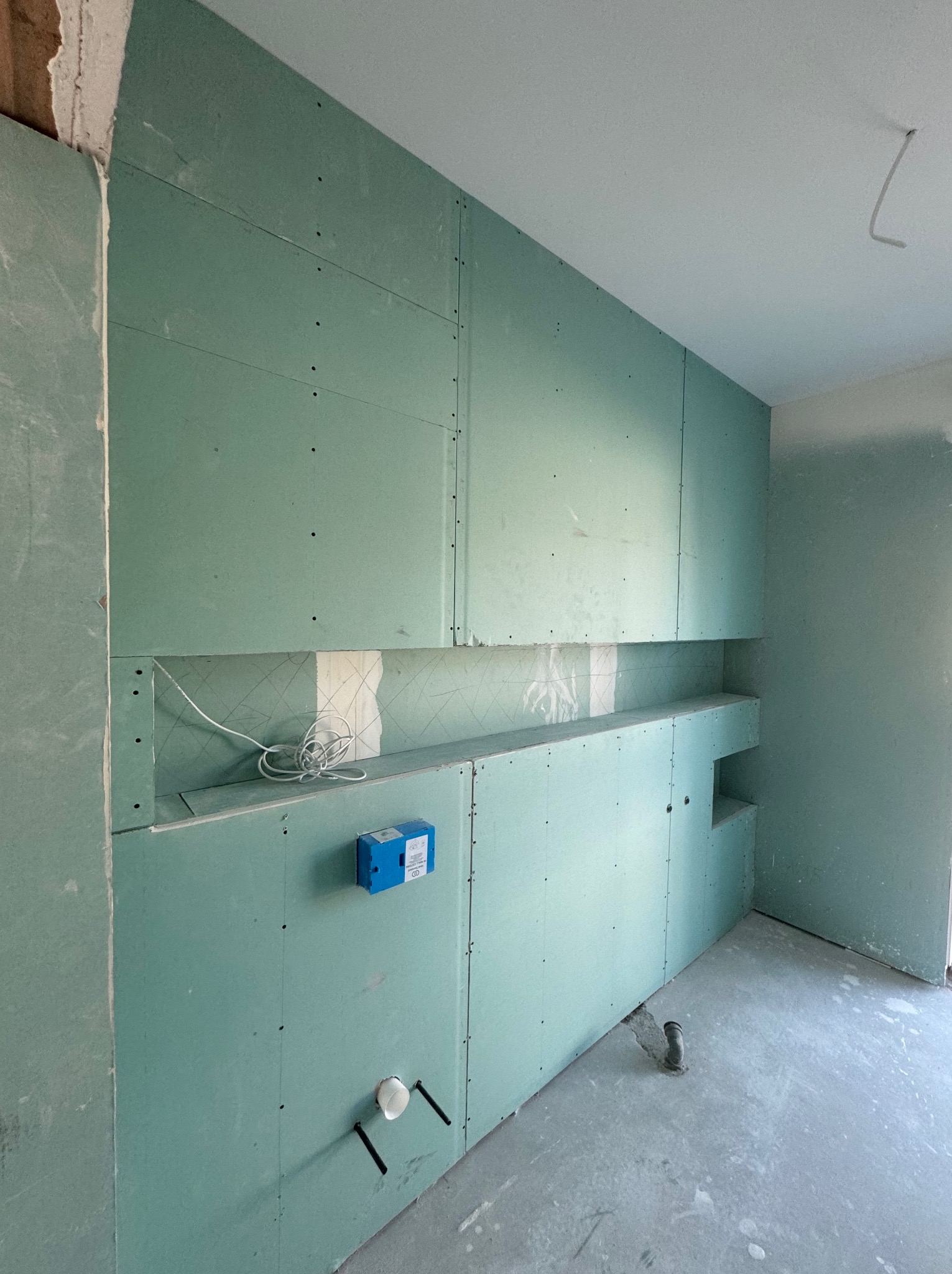 Ściana z zielonych płyt gipsowo-kartonowych z wnęką, widoczne przewody elektryczne, puszka instalacyjna i fragment instalacji hydraulicznej w surowym stanie.