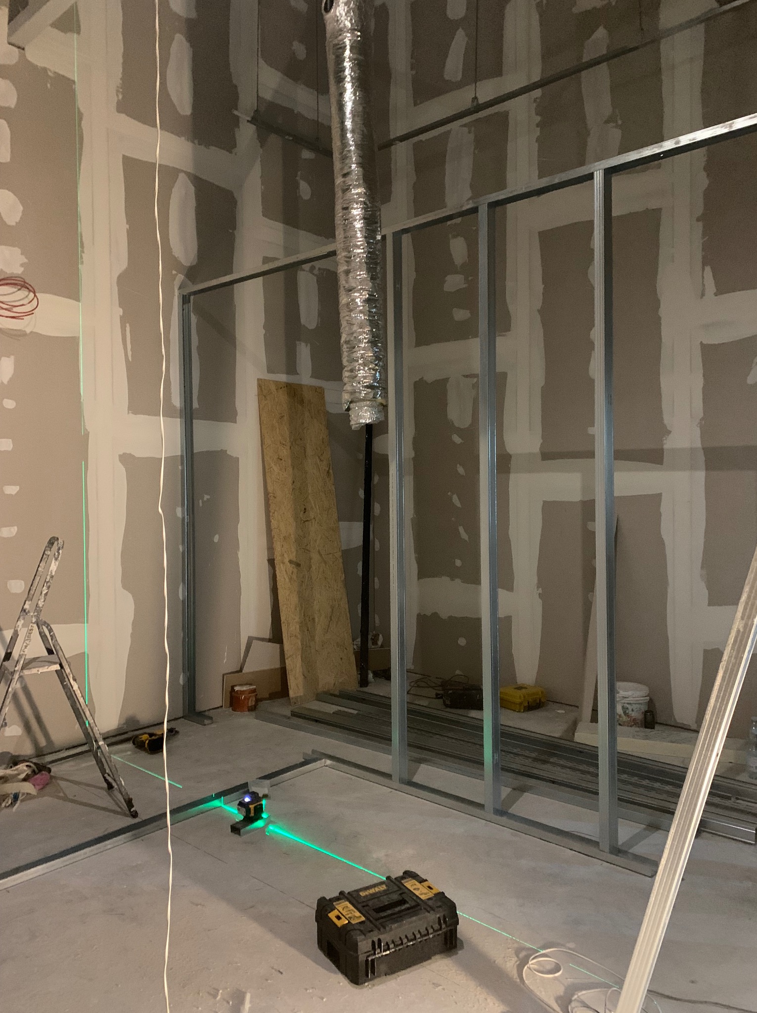 Wnętrze w trakcie montażu ścian działowych z profili metalowych, widoczne płyty kartonowo-gipsowe na ścianach i poziomica laserowa wyznaczająca linie, aluminiowy przewód wentylacyjny pod sufitem.