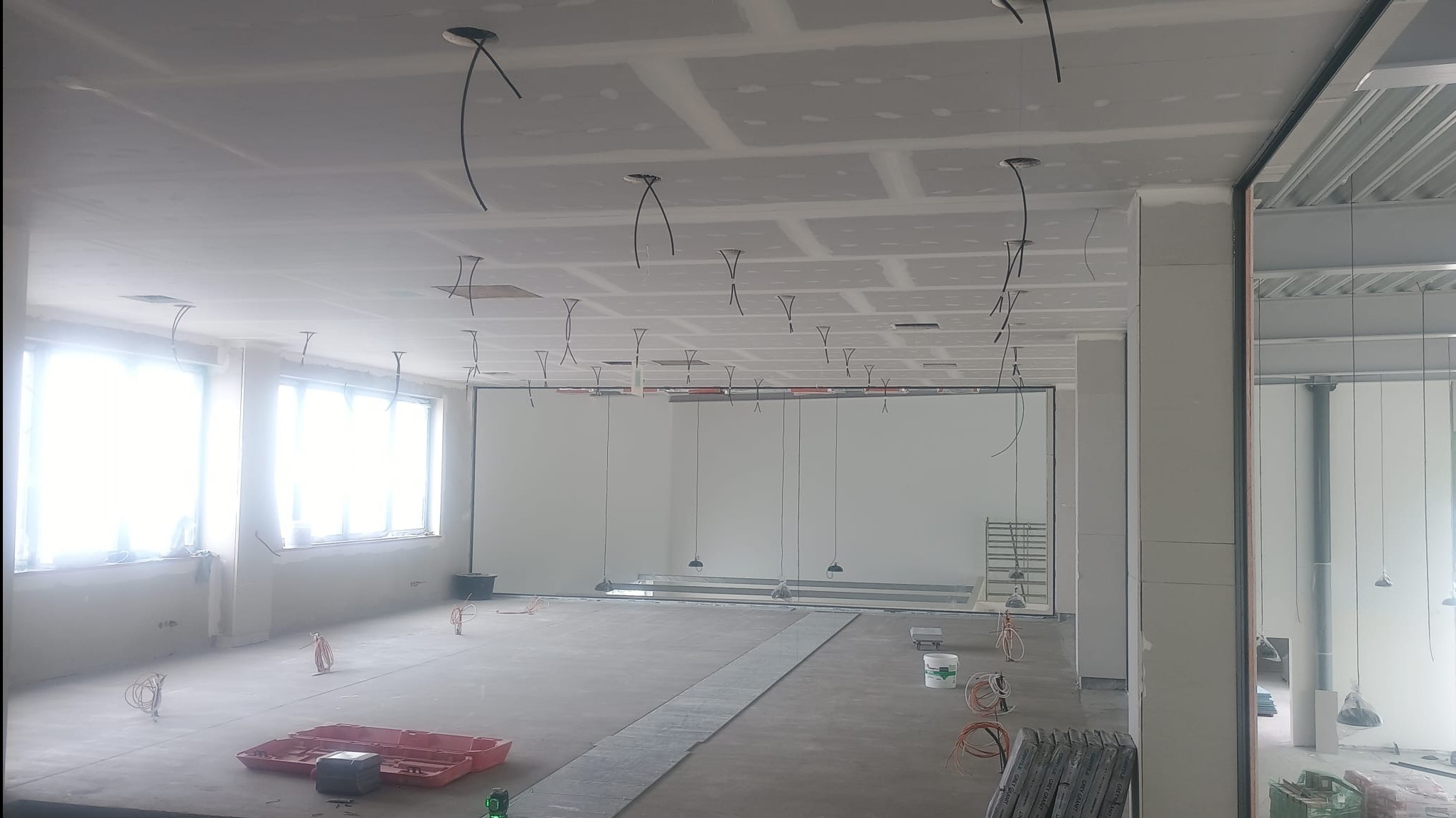 Wnętrze biura w trakcie prac wykończeniowych: zamontowane płyty karton-gips na suficie z wystającymi kablami, przygotowane podłoże pod posadzkę.