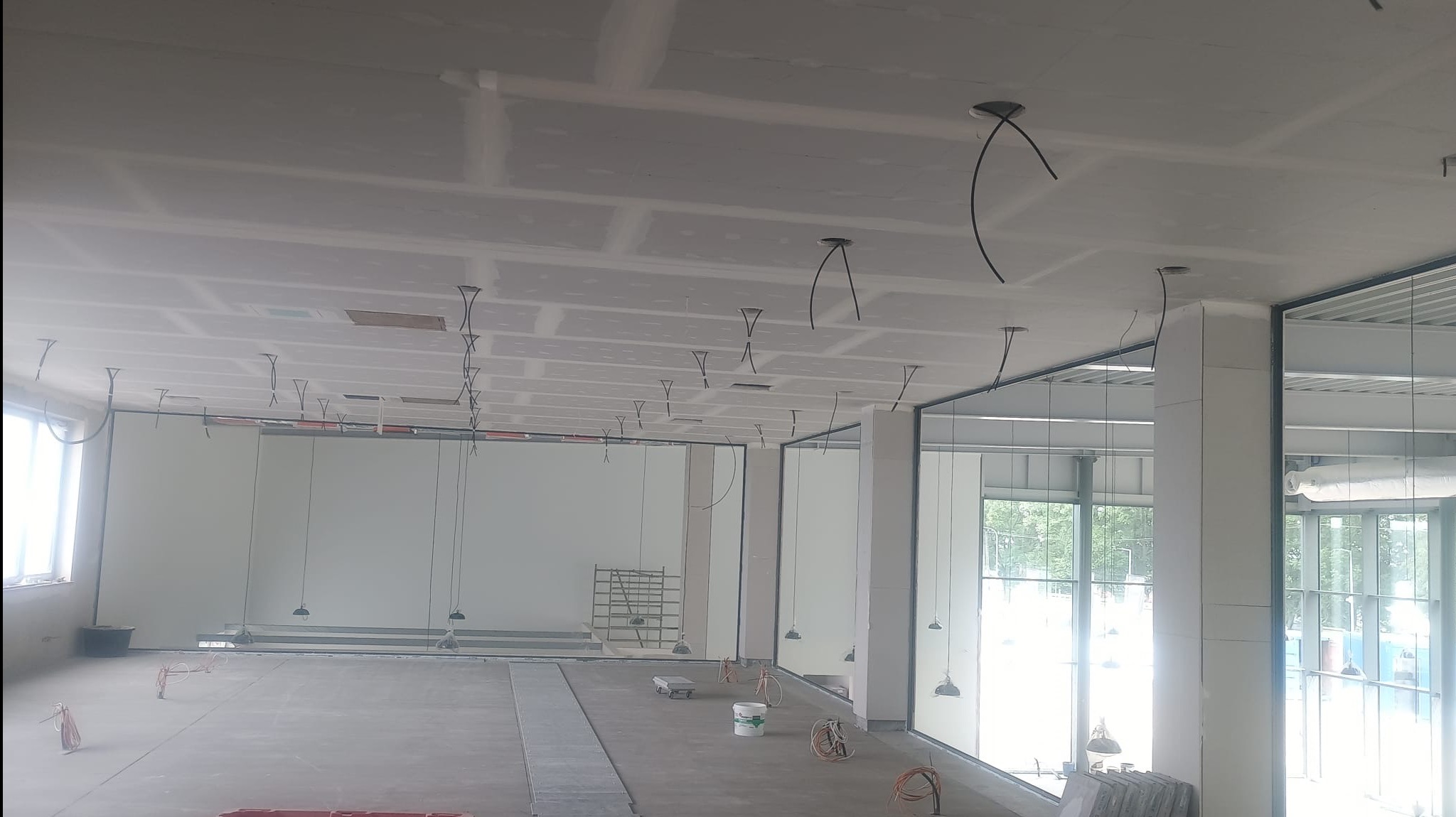 Sufit z płyt kartonowo-gipsowych w trakcie montażu, widoczne kable oświetleniowe i aluminiowe ramy okienne w jasnym, przestronnym wnętrzu komercyjnym.