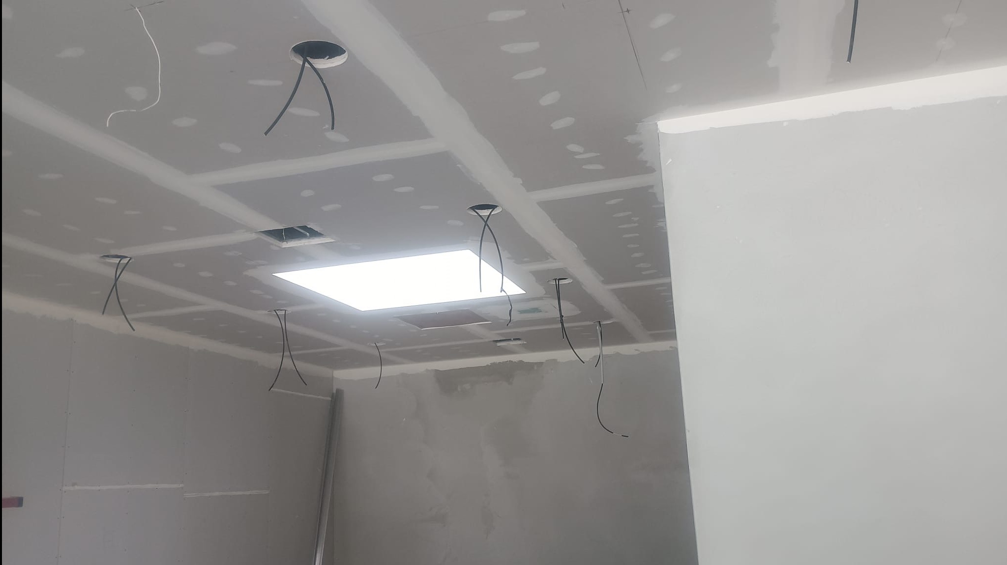 Sufit z płyt kartonowo-gipsowych w trakcie montażu, widoczne otwory na oświetlenie i wystające przewody elektryczne. Ściany przygotowane pod malowanie, surowe wykończenie.