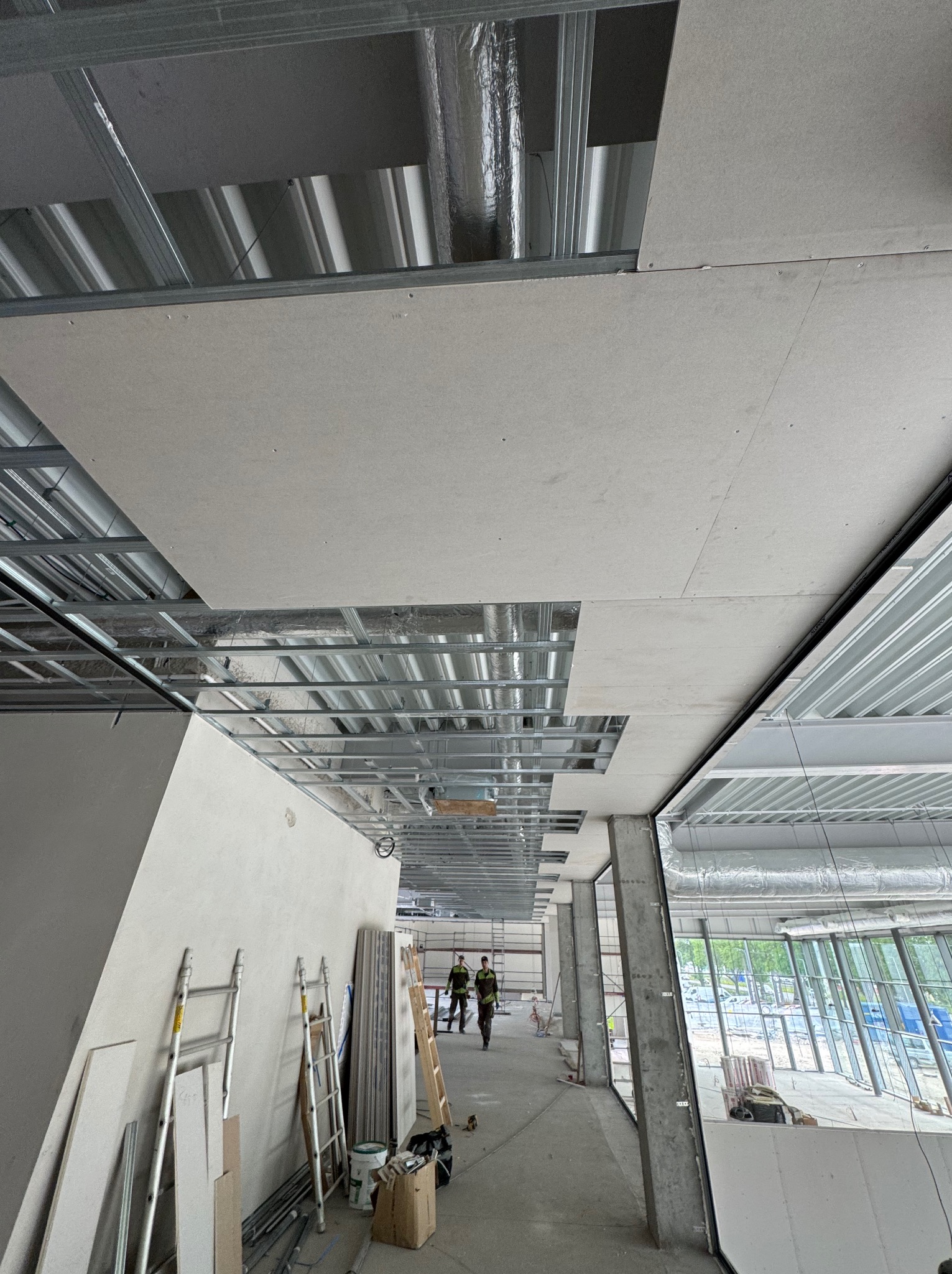 Montaż sufitu podwieszanego z płyt karton-gips na metalowej konstrukcji. Widoczne płyty, profile, drabiny i pracownicy w oddali. Etap prac wykończeniowych w budynku.