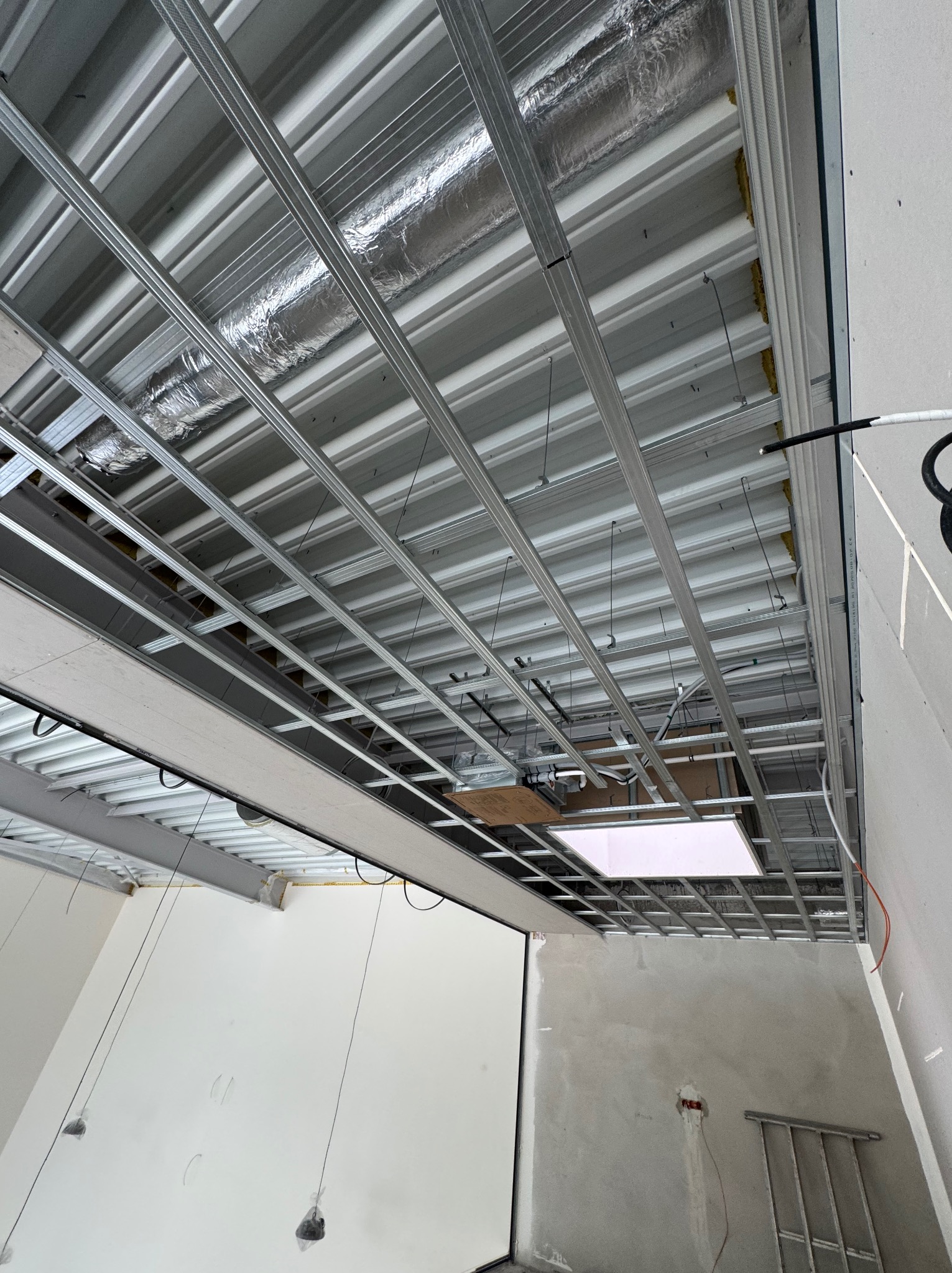 Metalowa konstrukcja sufitu podwieszanego z widoczną izolacją, instalacją elektryczną i elementami oświetlenia w surowym wnętrzu. Widok z dołu.