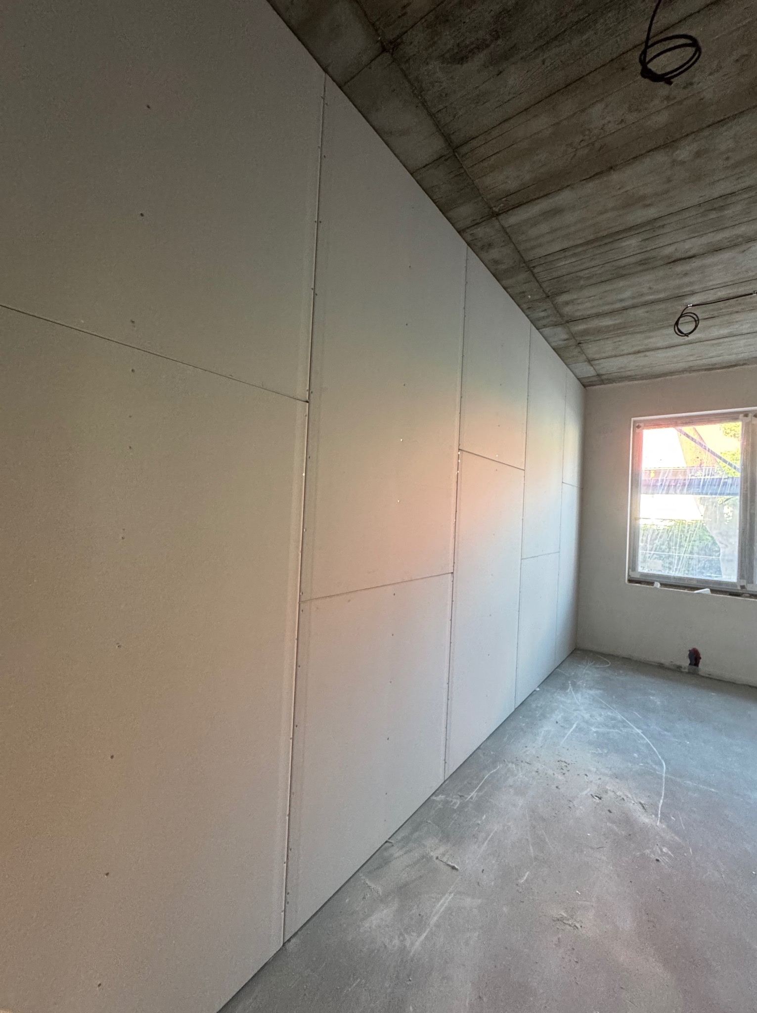 Montaż płyt na ścianie w surowym wnętrzu w Warszawie. Widoczne łączenia płyt i betonowy sufit. Okno z widokiem na zieleń.