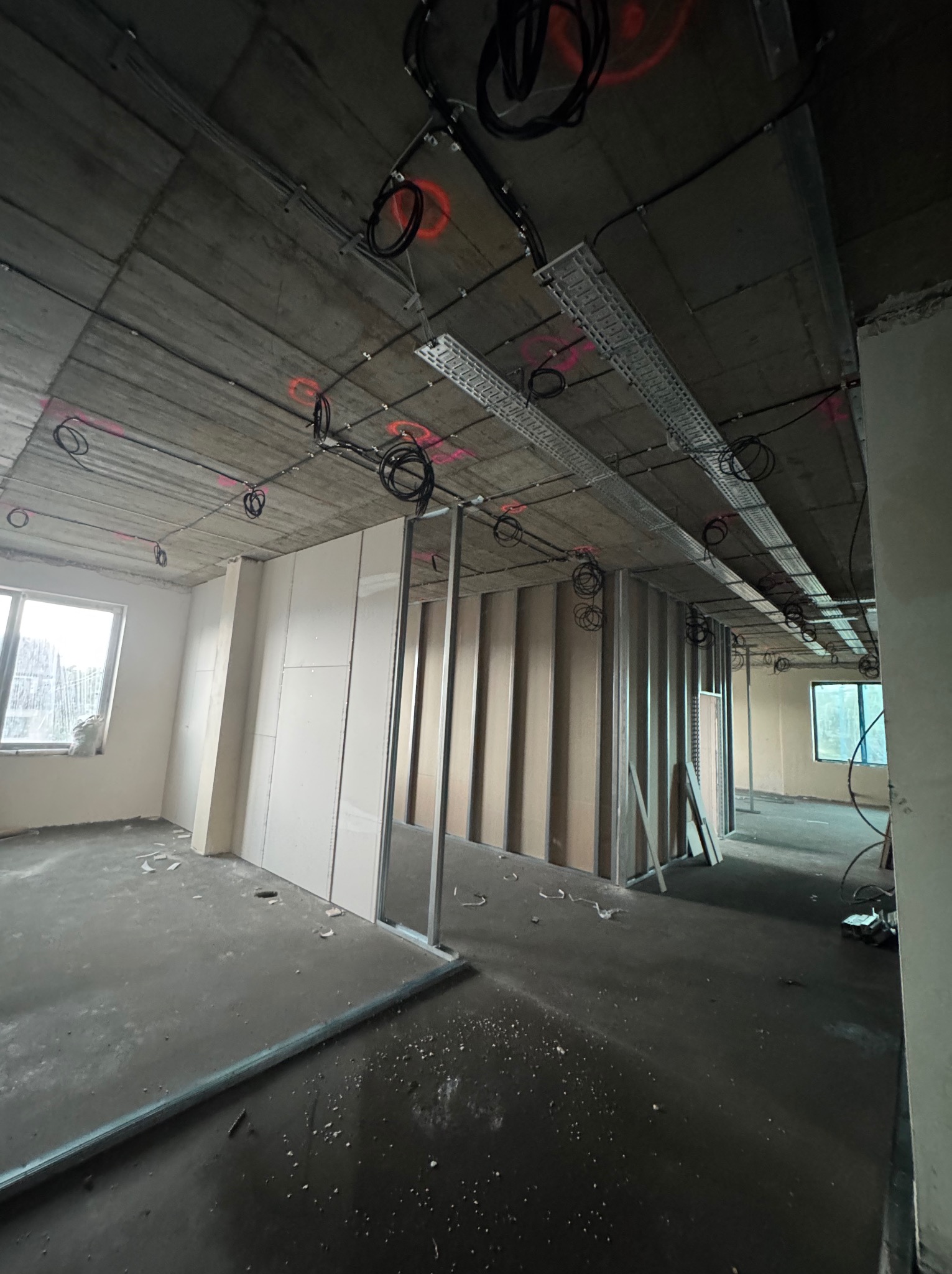 Wnętrze w trakcie prac wykończeniowych: montaż stelaża i płyt. Widoczne przewody, rury pod sufitem, okno z lewej strony. Surowy betonowy sufit z instalacjami.