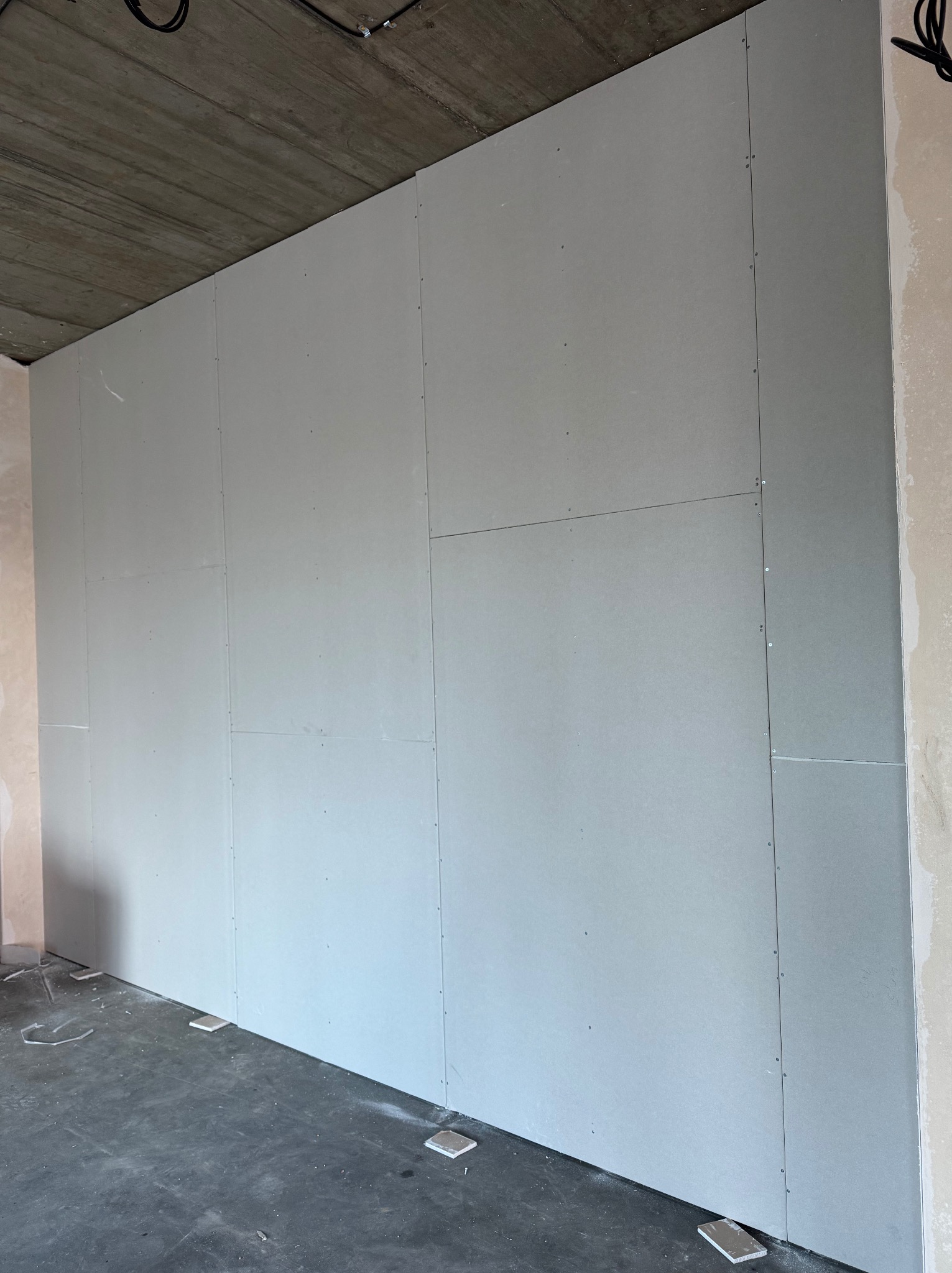 Ściana z płyt gipsowo-kartonowych, widoczne łączenia i wkręty, surowy betonowy sufit, przygotowanie do dalszych prac wykończeniowych, fragment podłogi.
